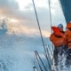 Sailors in orange gear navigate a yacht through rough seas, waves splashing, under a cloudy sky at sunset.