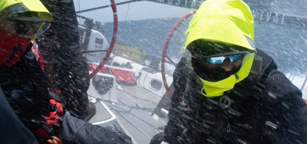 Sailors in yellow jackets navigate through rough seas during a sailing race, water splashing intensely on deck.