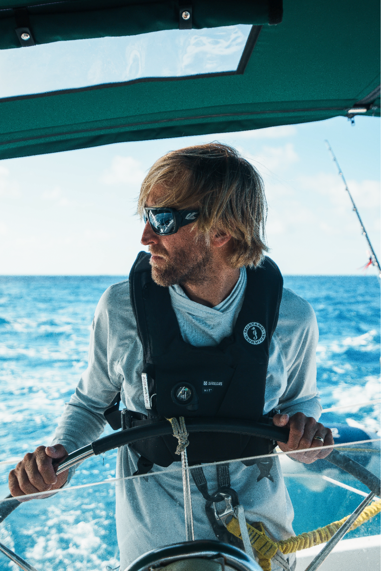 Man sailing a boat, wearing sunglasses and life vest, steering through blue ocean waters under clear skies.