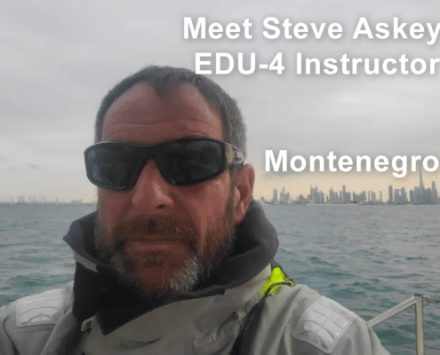 Steve Asley sailing in Montenegro, wearing sunglasses and sailing gear, city skyline in the background.
