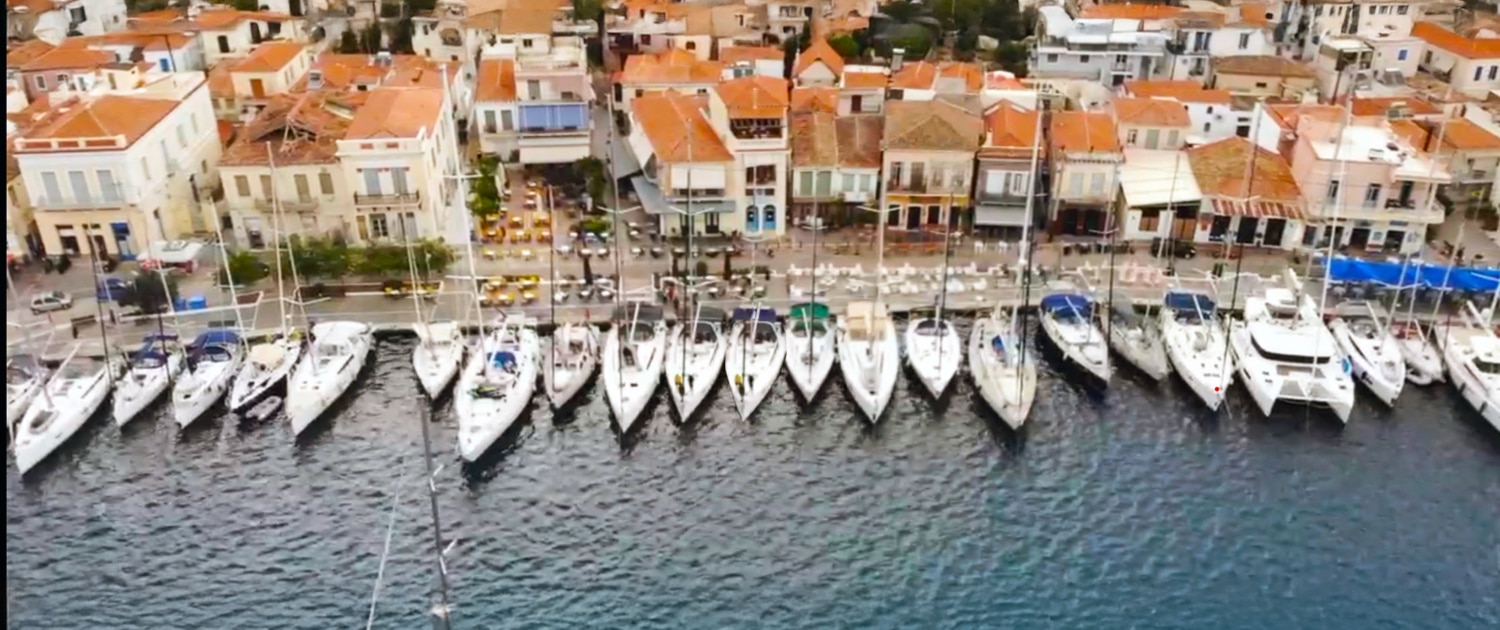 Aerial view of sailboats docked by a charming Mediterranean coastal village with orange-roofed buildings.