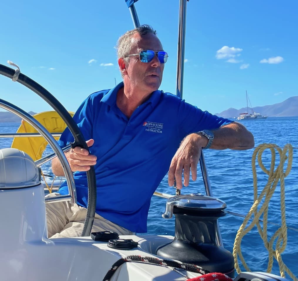 Screenshot_20230323_202525_Email Man steering sailboat on calm sea, wearing blue shirt and sunglasses, with mountains in the background.