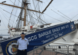 USCG Barque Eagle docked with uniformed person, banner reads America's Tall Ship. Sailboat in background.