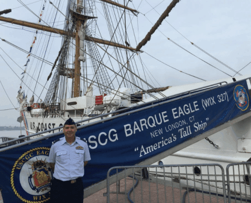 USCG Barque Eagle docked with uniformed person, banner reads America's Tall Ship. Sailboat in background.