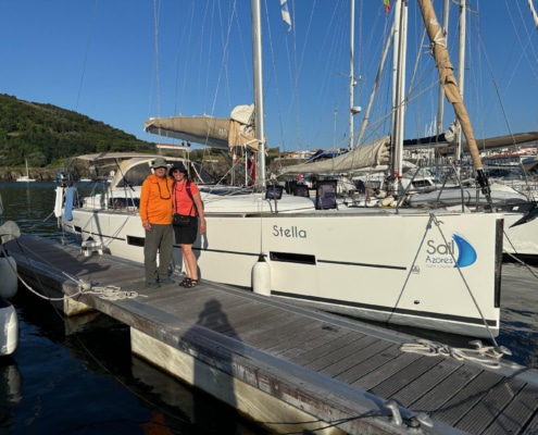 Dave Stern Couple posing beside Stella sailboat at a marina, with clear blue sky and lush hillside in the background.