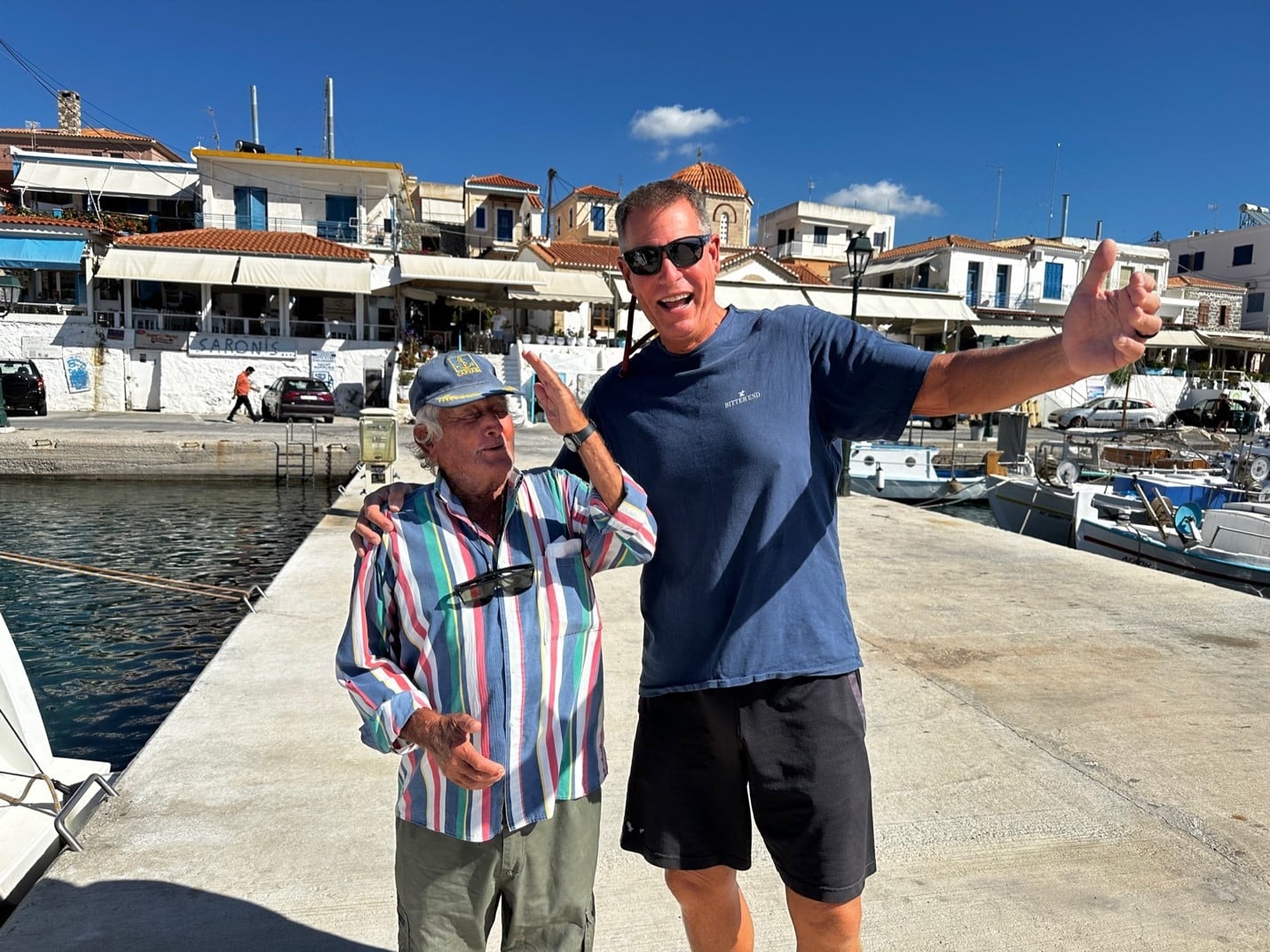 AEGINA Two men happily pose near a marina with boats and charming seaside buildings in the background.