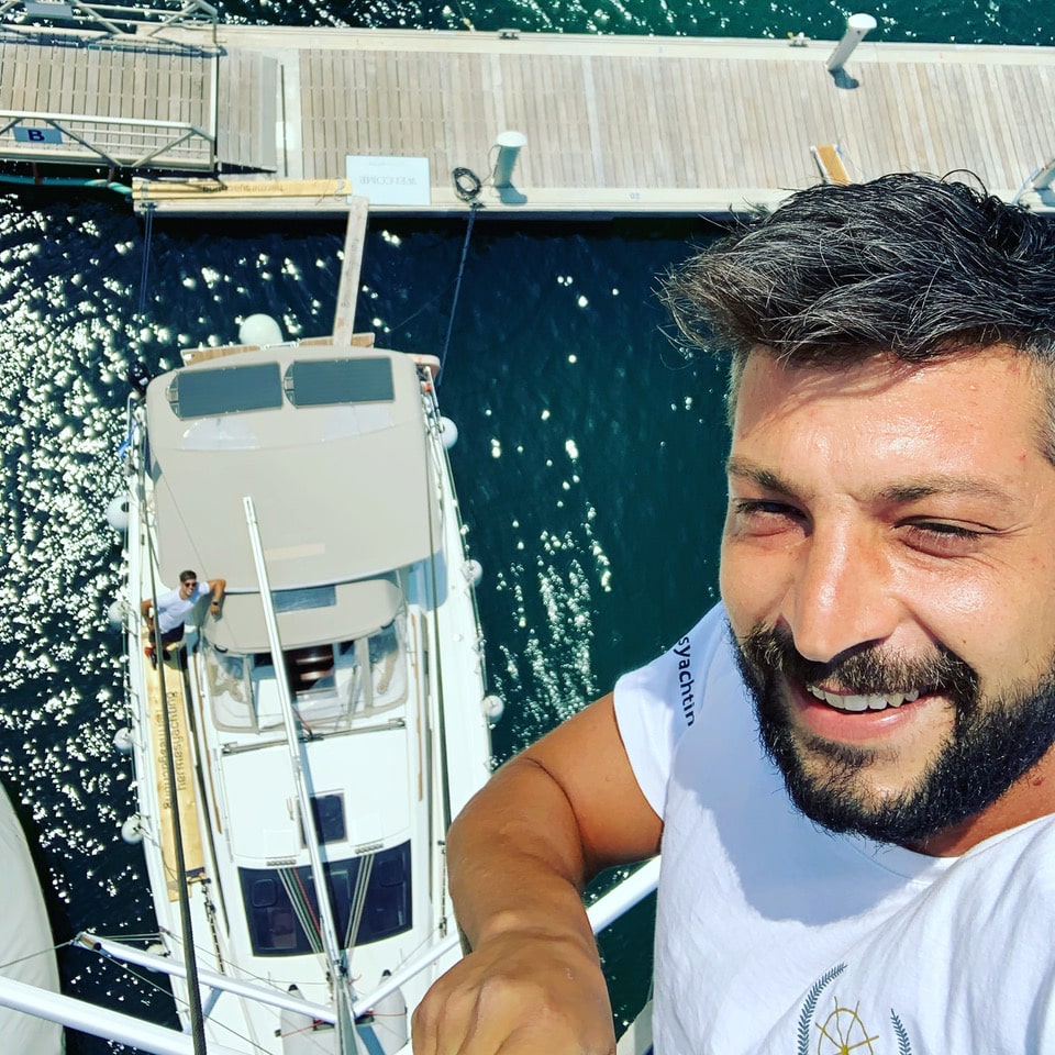 1CABEE75-3BA2-46AB-ABC6-F3C550161518 Smiling man atop sailboat mast above marina, capturing thrilling view of yacht and sparkling water below.