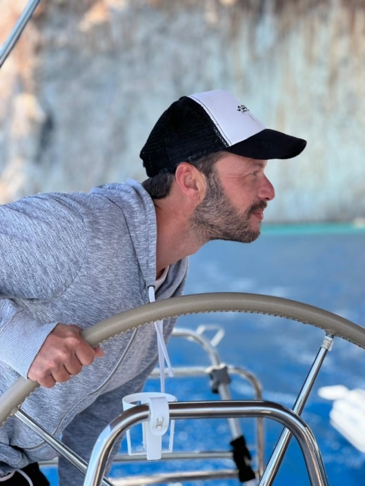 IMG-20230528-WA0019 Man steering sailboat, focused and determined in casual attire, navigating calm blue waters with rocky background scenery.