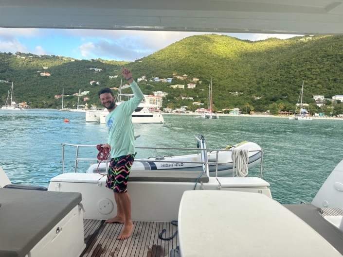 IMG-20240229-WA0016 Man happily holding rope on sailboat deck with lush green hills and sailboats in the background.