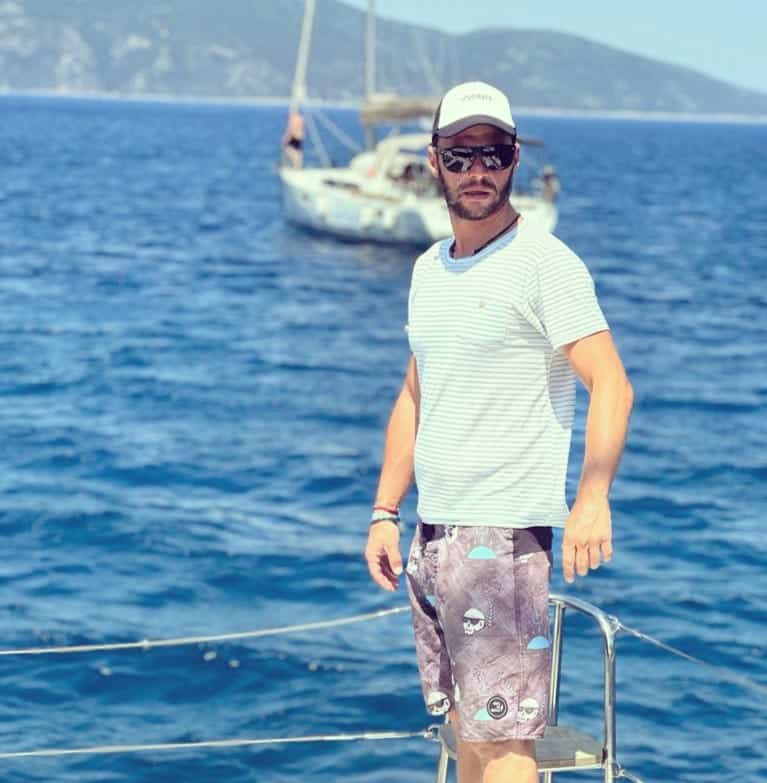 Man enjoying a sunny day on a sailboat with scenic ocean and distant hills in the background.