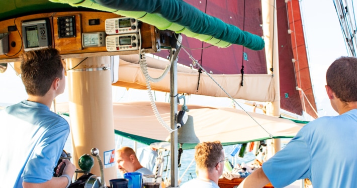 Scouts steering a sailboat with colorful sails Sea Scout Crew navigating a sailing ship with red sails, focusing on the helm and gauges, under clear skies.