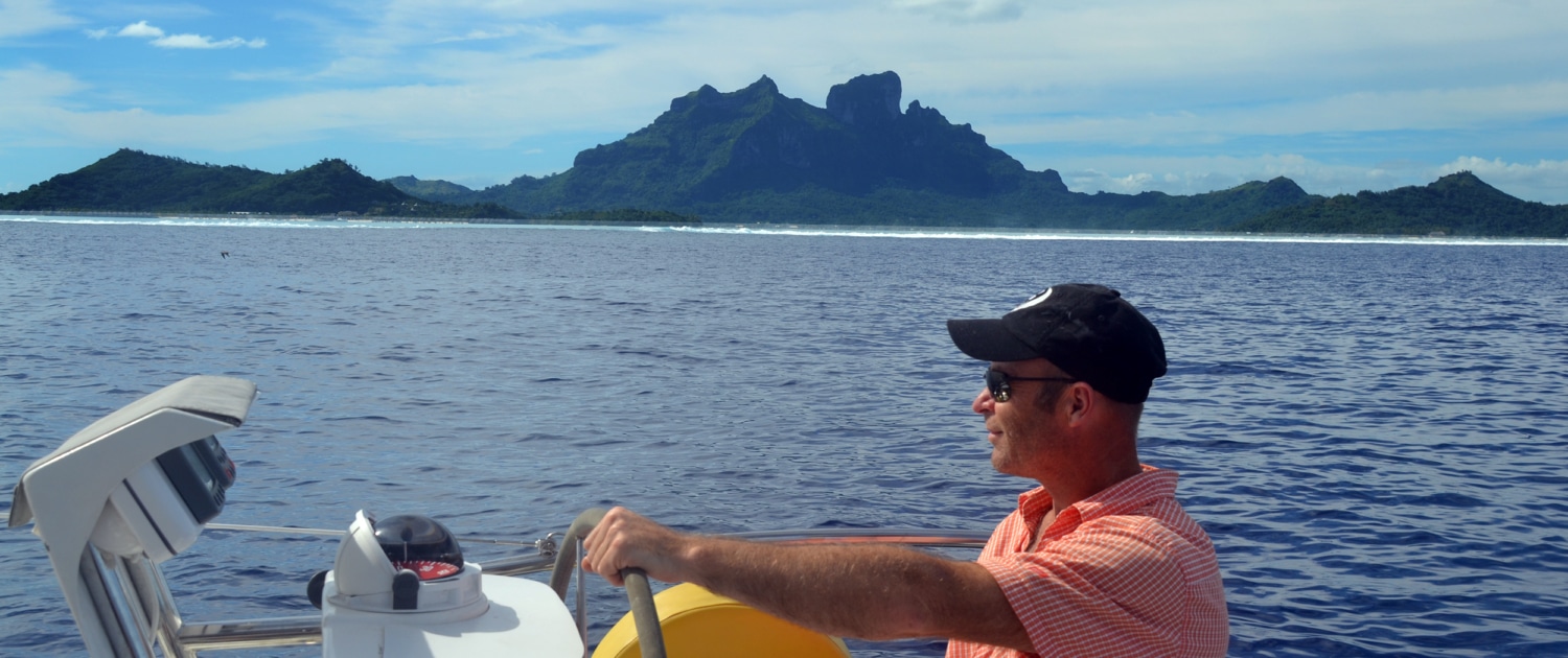 Man steering sailboat with island backdrop, wearing a cap and sunglasses, sunny day on the ocean.