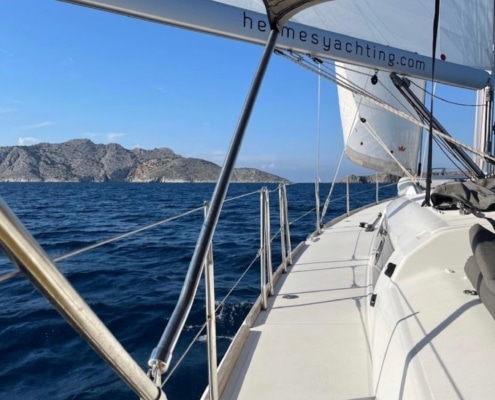 George Sakkas Sailing yacht on a clear blue sea with distant rocky coastline in view under a sunny sky; serene maritime adventure.