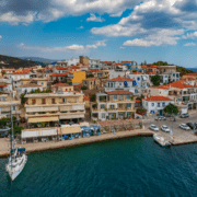 nauticed-2026-09-athens-flotilla-blog-49-portoheli05 Scenic Greek coastal town with colorful buildings, waterfront cafes, clear blue skies, and anchored sailboat in harbor.
