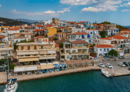 Scenic Greek coastal town with colorful buildings, waterfront cafes, clear blue skies, and anchored sailboat in harbor.