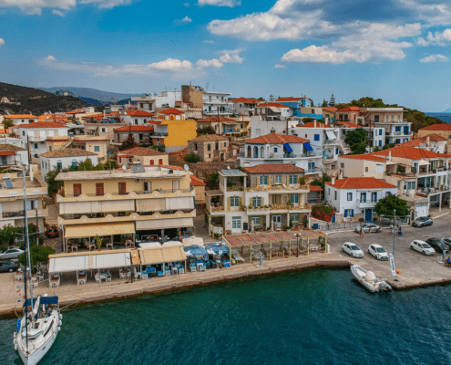 Scenic Greek coastal town with colorful buildings, waterfront cafes, clear blue skies, and anchored sailboat in harbor.