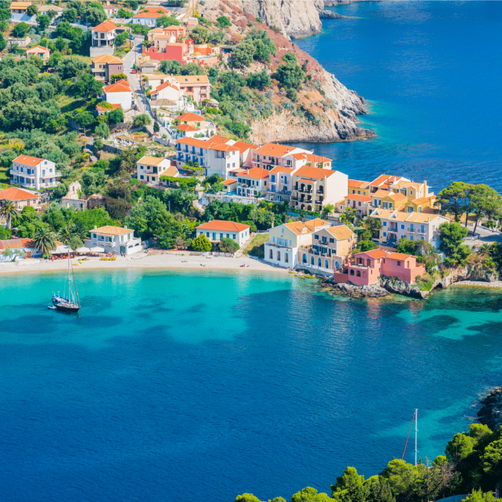 blog-below-deck-destinations-1-Ionians Scenic coastal village with colorful houses, turquoise waters, and anchored sailboats, nestled in lush greenery.