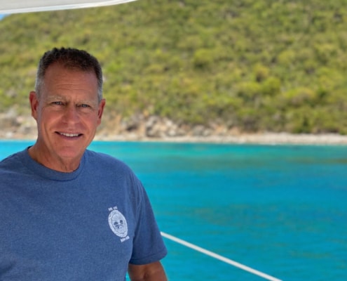 Bill Ibsen Smiling man on a sailboat with turquoise waters and lush island in the background. Perfect day for sailing adventure!