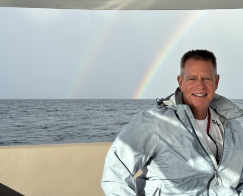 Bill Ibsen Man smiling on a boat at sea with a vibrant rainbow in the background.