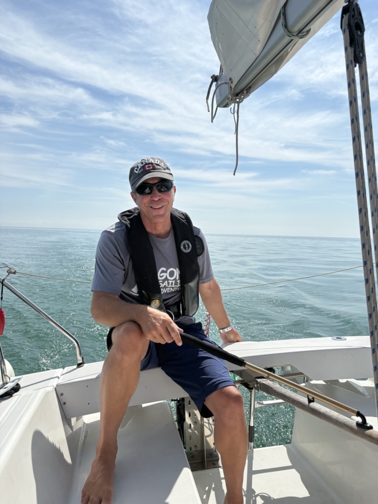 Robert Frigault Man smiling while steering a sailboat on a sunny day at sea, wearing sunglasses and a cap.
