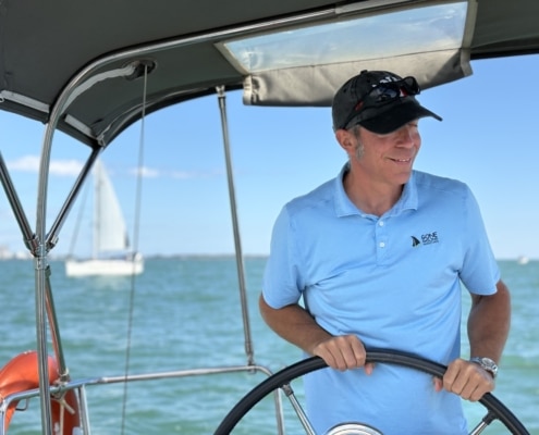 Robert Frigault Man steering a sailboat under a canopy on a sunny day, with sailboat and ocean in the background.