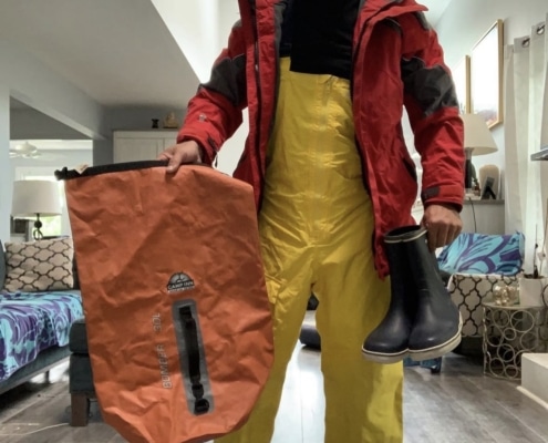 Robert Frigault Man in sailing gear with waterproof bag and boots, preparing for a nautical adventure indoors.