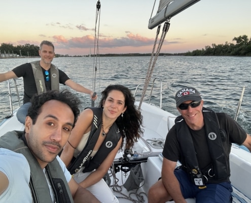 Robert Frigault Group enjoying sunset sailing adventure on a calm lake, wearing life jackets and smiling on a sailboat.