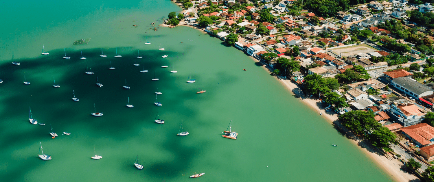 Aerial view of sailboats anchored in turquoise waters near a coastal town with colorful homes and lush greenery.