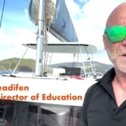 A person on a sailboat with lush islands and boats in the background, wearing green sunglasses and a NauticEd shirt.