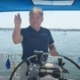 Man steering sailboat with confidence on a sunny day, showcasing sailing gear and open water scenery.
